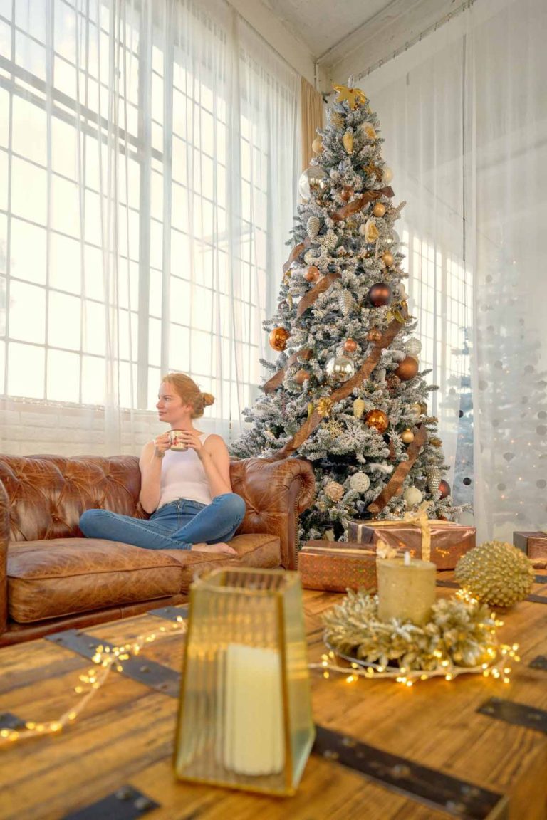 Woman relaxing on a sofa with a cup of tea in a cozy living room beside a decorated Christmas tree and festive holiday decor
